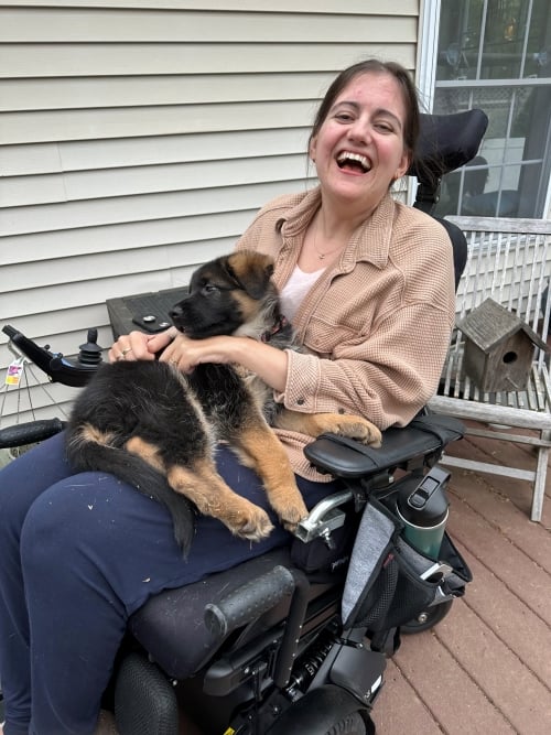 A woman in a black power wheelchair wearing a beige coat and holding a puppy. She is laughing.