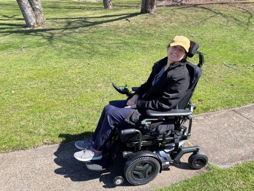 A woman smiling in a black power wheelchair on a park sidewalk