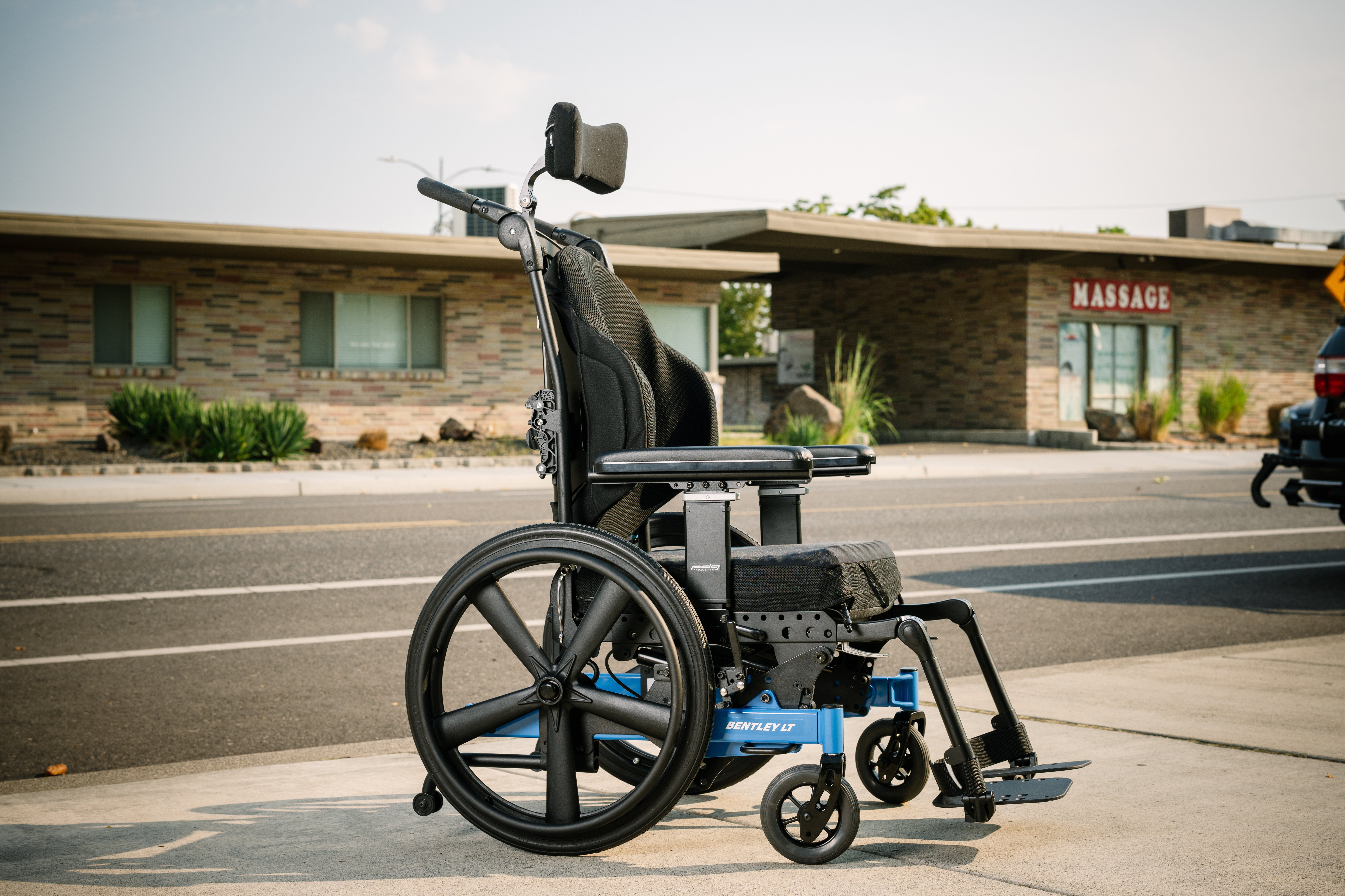 A blue and black PDG Bentley LT wheelchair sits on the sidewalk in front of a small road